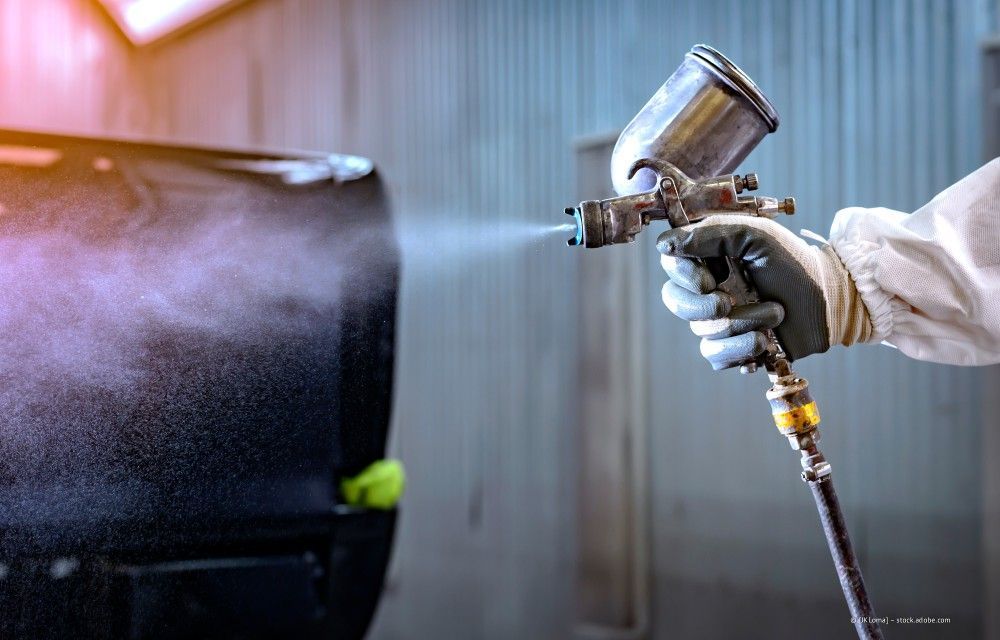Eine Person besprüht in einer Industrieumgebung ein schwarzes Fass mit einer silbernen Spritzpistole.