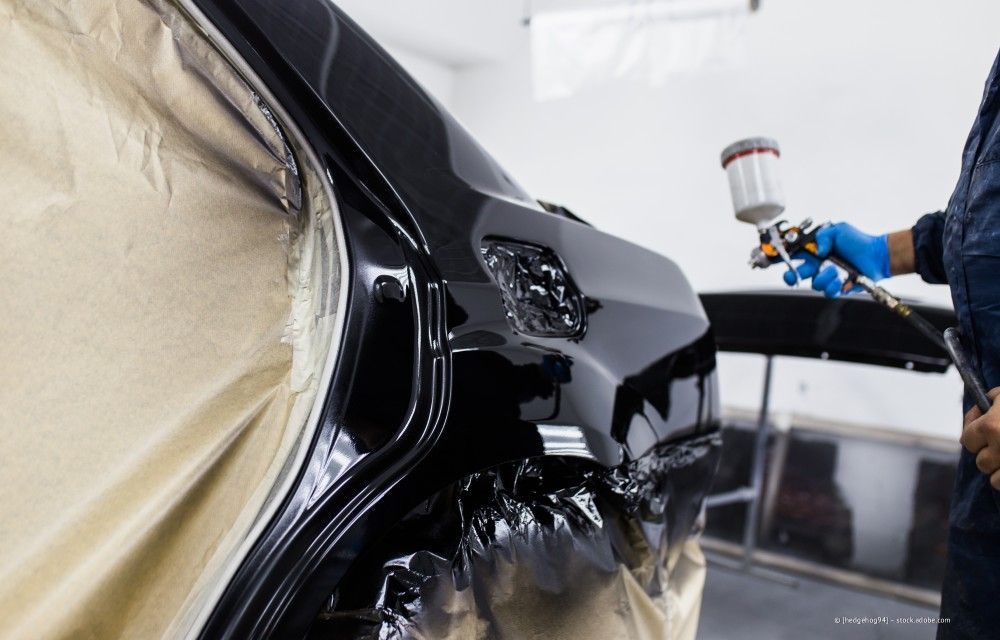 Eine schwarze Autokarosserie wird in einer Werkstatt lackiert; Teile des Autos sind abgeklebt, und ein Lackierer benutzt eine Spritzpistole.