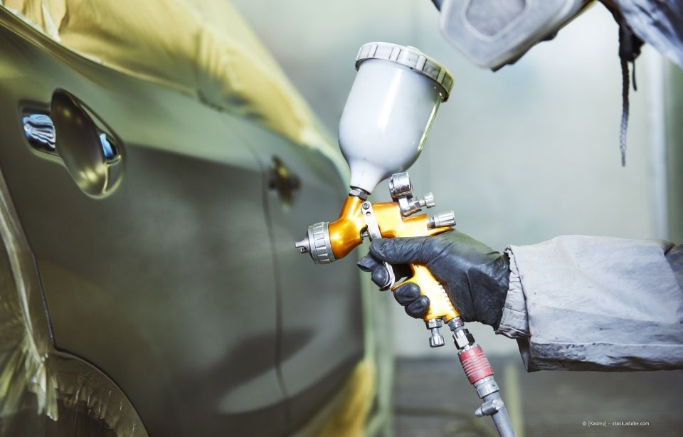 Ein Arbeiter lackiert in einer Werkstatt eine Autotür mit einer Farbspritzpistole und weißen Handschuhen.