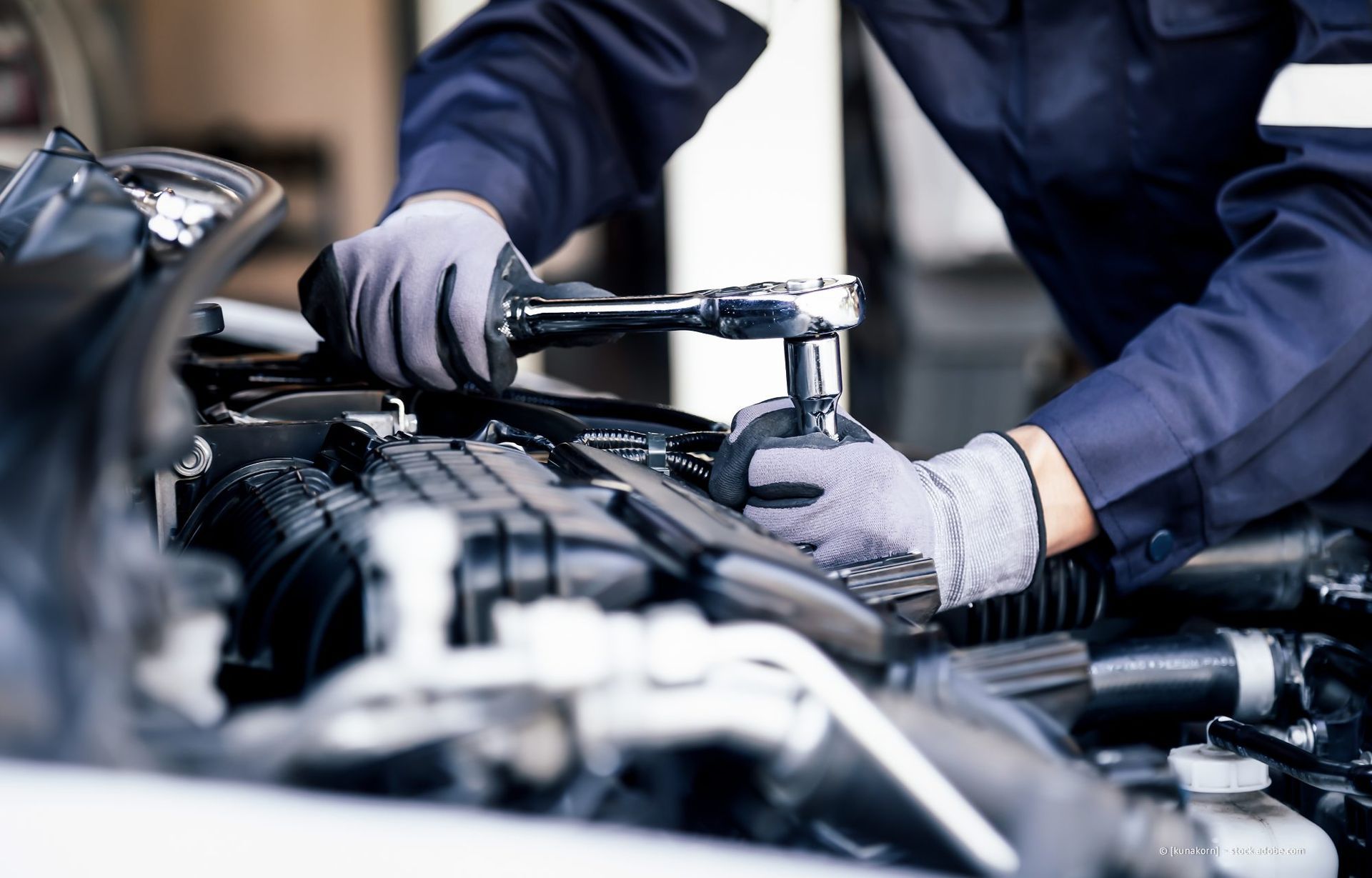 Ein Mechaniker mit Handschuhen zieht Motorteile unter einer geöffneten Motorhaube fest.