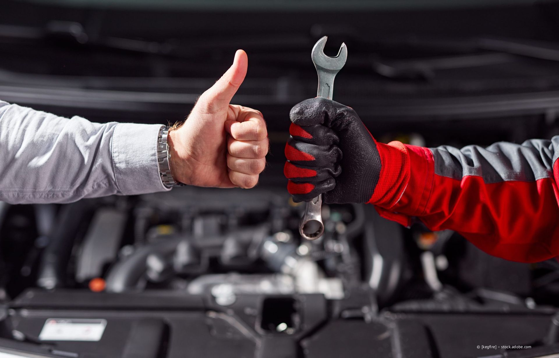 Zwei Mechaniker tauschen einen Daumen hoch und einen Schraubenschlüssel über einem offenen Automotor aus.