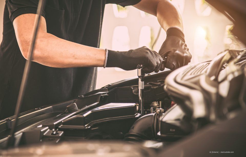 Ein Mechaniker arbeitet mit behandschuhten Händen unter einer Motorhaube in einer Garage.