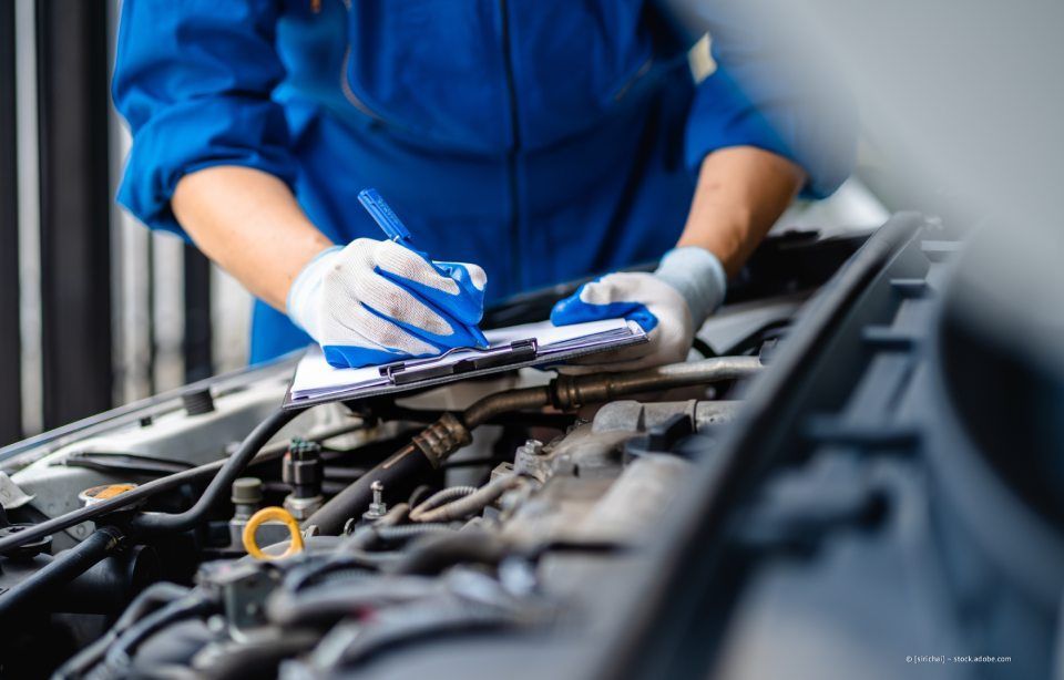 Mechaniker in blauer Arbeitskleidung überprüft mit Klemmbrett und Handschuhen Motorkomponenten.