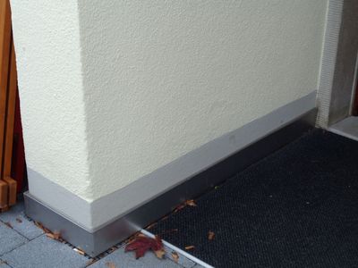 Person mit Handschuhen, die eine weiße Dachrinne an einem Backsteingebäude installiert, auf dem ein grünes Dach sichtbar ist.