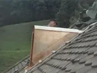Graues Haus mit dunklem Dach und zwei Dachgauben, umgeben von grünen Bäumen.