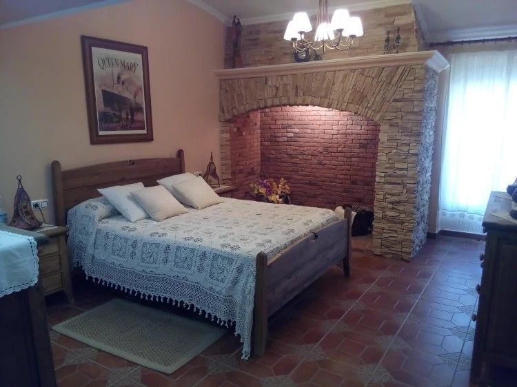 Dormitorio con cama de madera, chimenea de ladrillo, suelo de terracota y obras de arte enmarcadas.