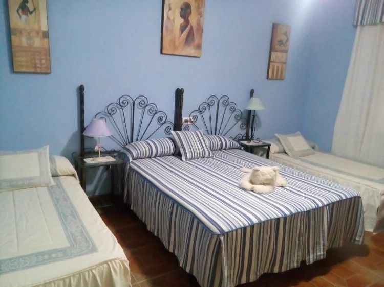 Dormitorio con tres camas; paredes azules, ropa de cama a rayas, cabeceros de metal, obras de arte, mesitas de noche.