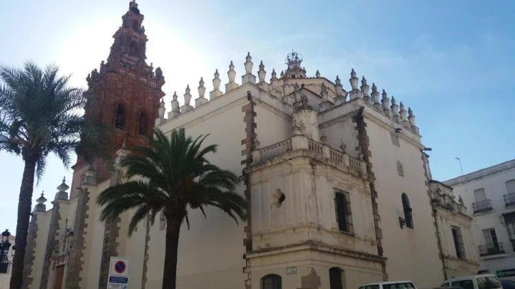 Edificio de la iglesia con fachada blanca adornada y torre roja, palmeras en el frente bajo un cielo azul.