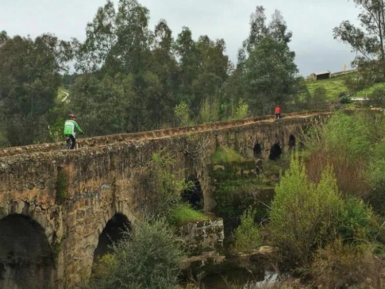 Dos ciclistas en un viejo puente de piedra sobre un río, rodeados de árboles. Día nublado.