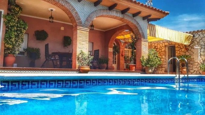 Piscina de agua azul frente a una casa estilo español con arcos, plantas y patio.
