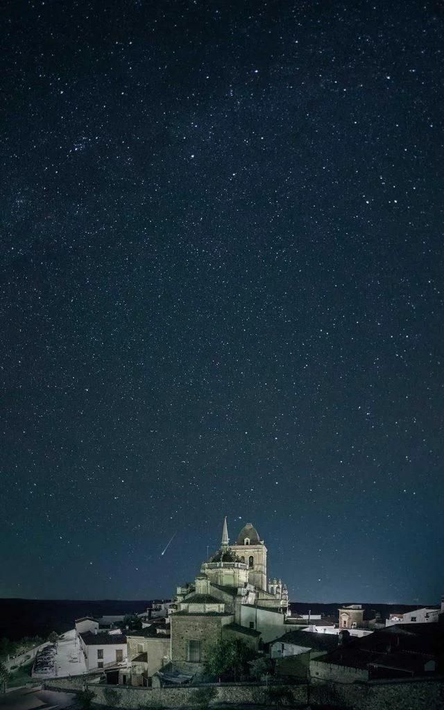 Cielo nocturno sobre una iglesia de piedra con numerosas estrellas visibles, posiblemente en un pueblo rural.