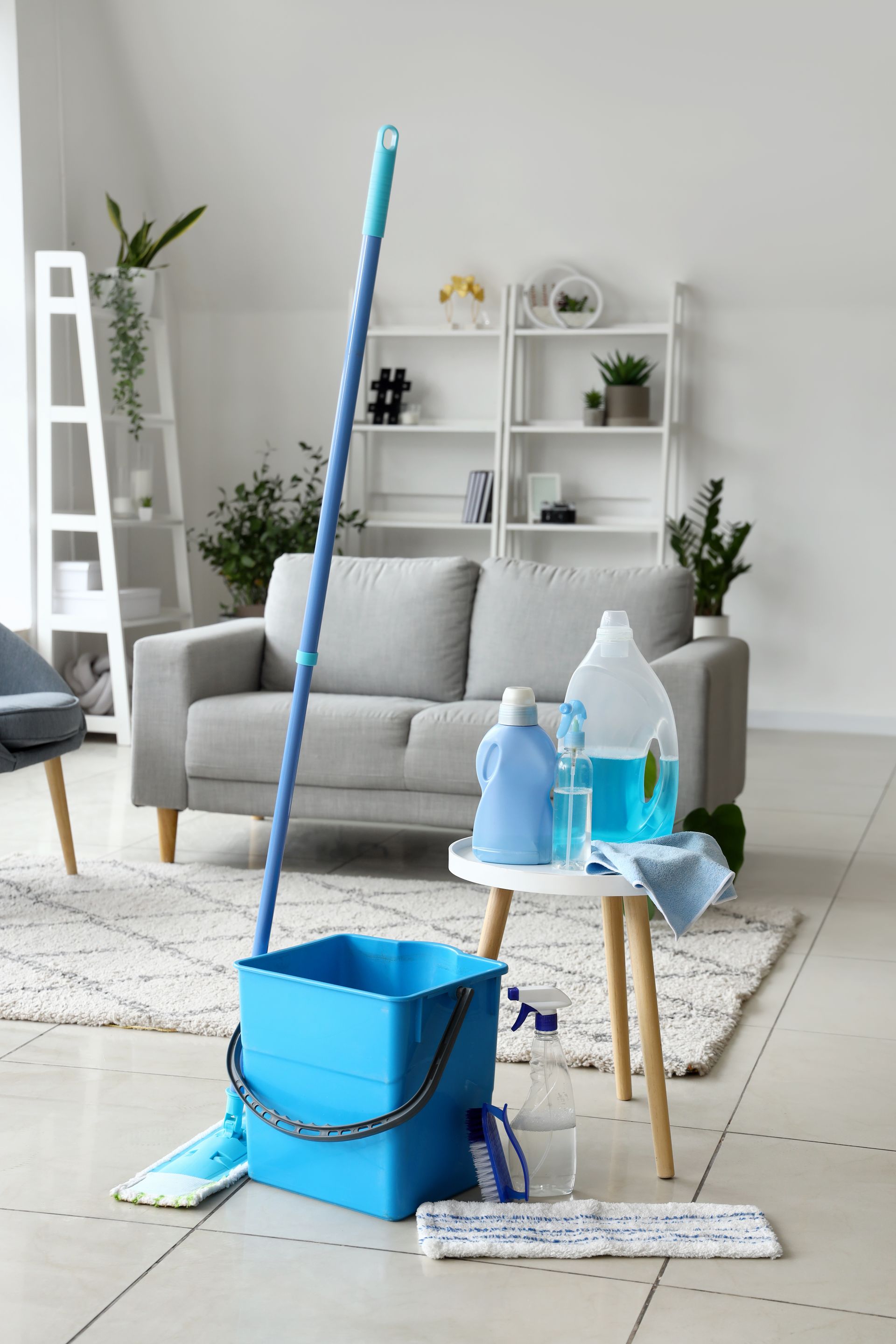 Des produits de nettoyage bleus, dont une serpillière et un seau, sur un sol carrelé dans un salon.