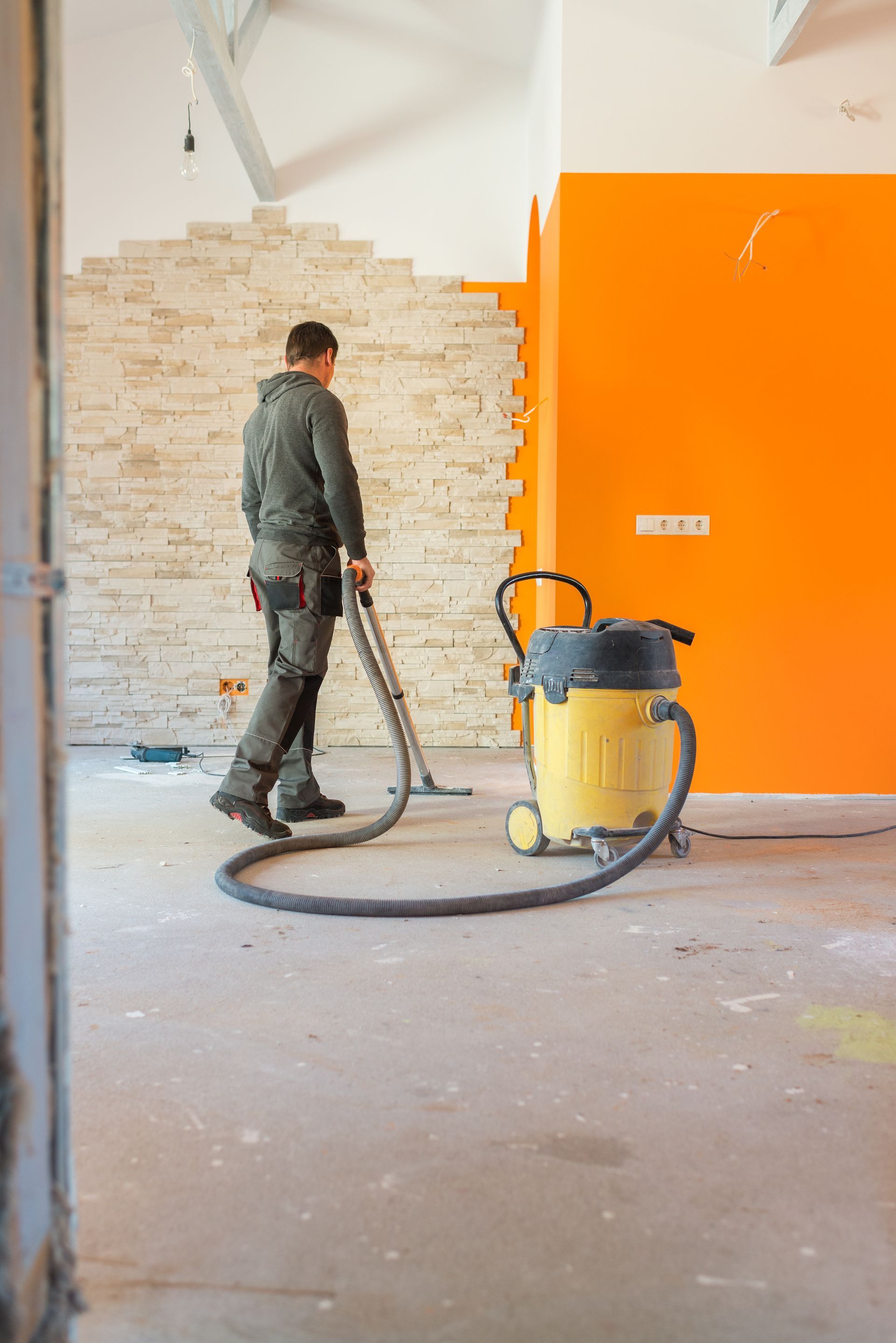 Une personne passe l'aspirateur dans une pièce aux murs orange et en briques ; un aspirateur jaune est visible.