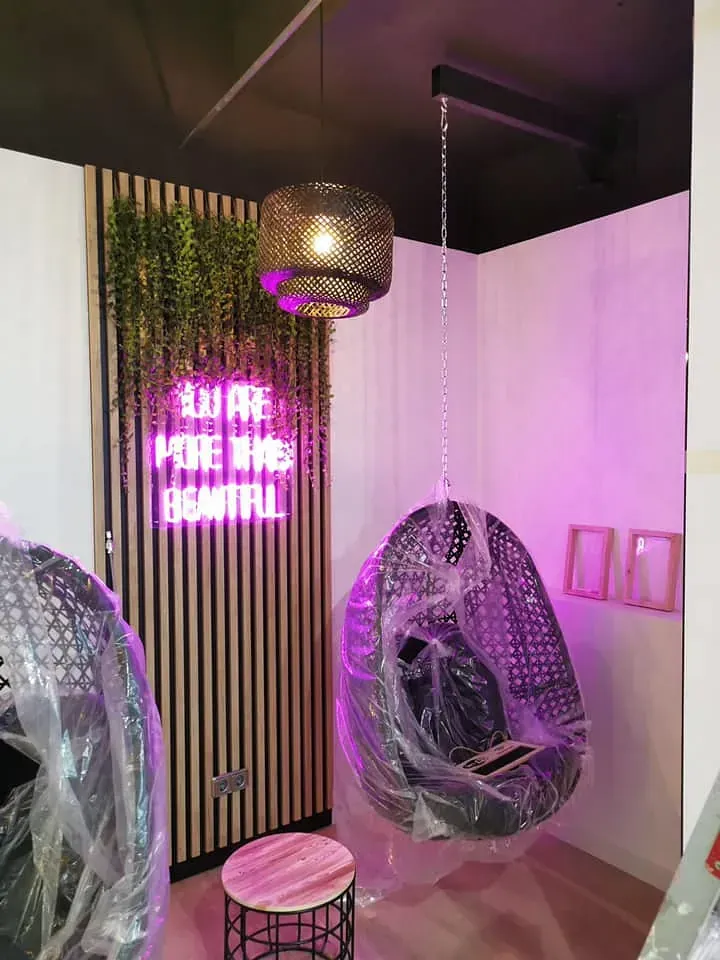 Una habitación con un letrero de neón que dice "ERES MÁS QUE HERMOSA", una silla colgante y una lámpara colgante.
