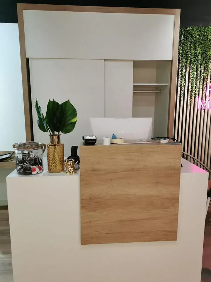 Mostrador de recepción con panel imitación madera, paredes blancas y elementos decorativos: hojas, jarrón, jarras y estantes en un entorno luminoso.