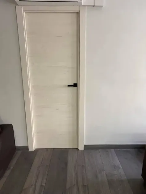 Puerta blanca con manija negra, colocada en un marco blanco contra una pared blanca, sobre un piso con apariencia de madera.