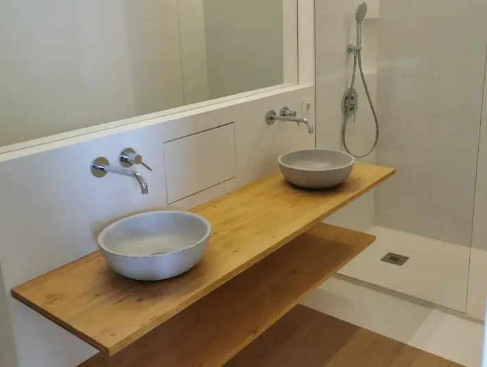 Baño con estantes de madera, dos lavabos y ducha a ras de suelo. Paredes blancas y suelo de madera marrón claro.