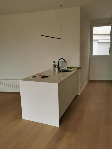 Isla de cocina blanca con fregadero, grifo negro y luz de techo. Suelos y paredes de madera clara.