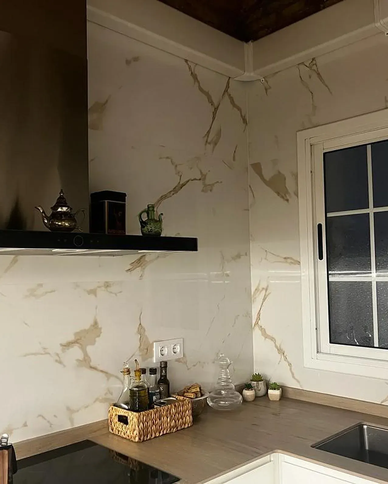 Cocina con paredes efecto mármol, campana extractora oscura, ventana y encimera. Elementos decorativos en estantes y encimeras.
