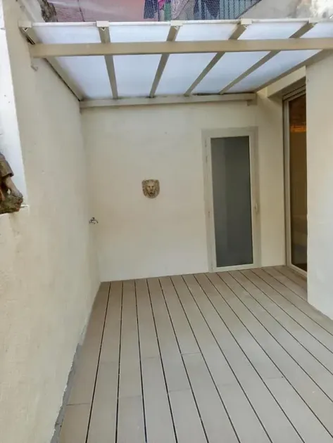 Pequeño patio con tablones de madera claros. Paredes blancas, puerta de cristal y techo translúcido.