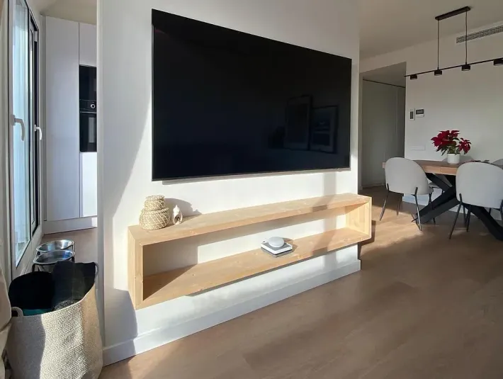 Sala de estar con TV montada, estante flotante y comedor, bañado por la luz del sol.