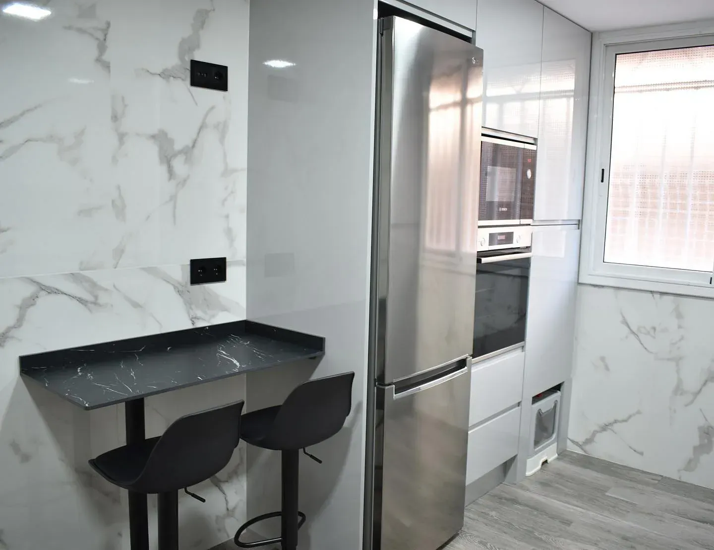 Cocina moderna con paredes efecto mármol, frigorífico de acero inoxidable, taburetes negros y una pequeña barra.