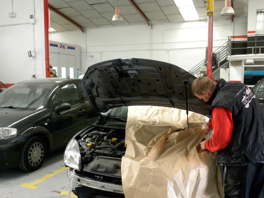 Un hombre repara un coche en un taller, enmascarando la parte delantera. Se ve otro coche cerca.
