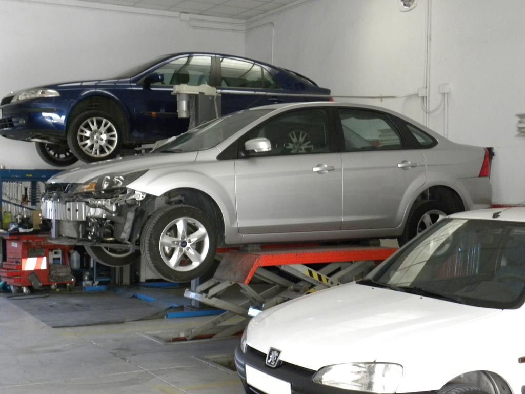 Coches en un garaje, uno de ellos subido a un elevador. El coche plateado presenta daños en la parte delantera. El coche azul está en un elevador superior. El coche blanco aparece en primer plano.