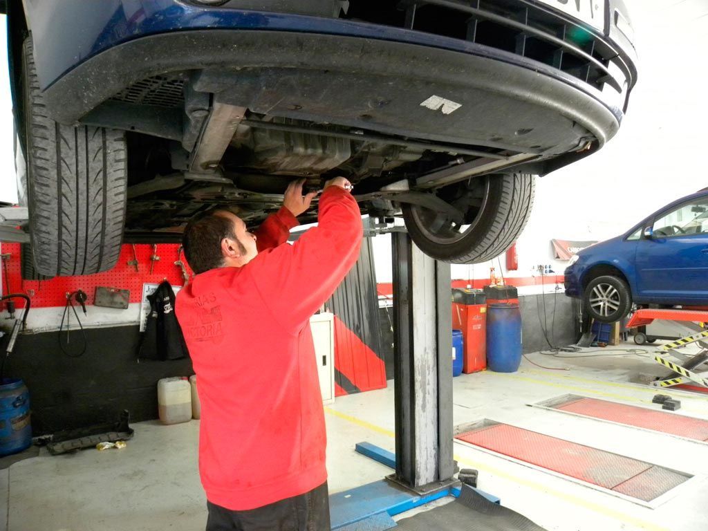 Mecánico con camisa roja trabajando debajo de un coche azul levantado mediante un polipasto en un garaje.