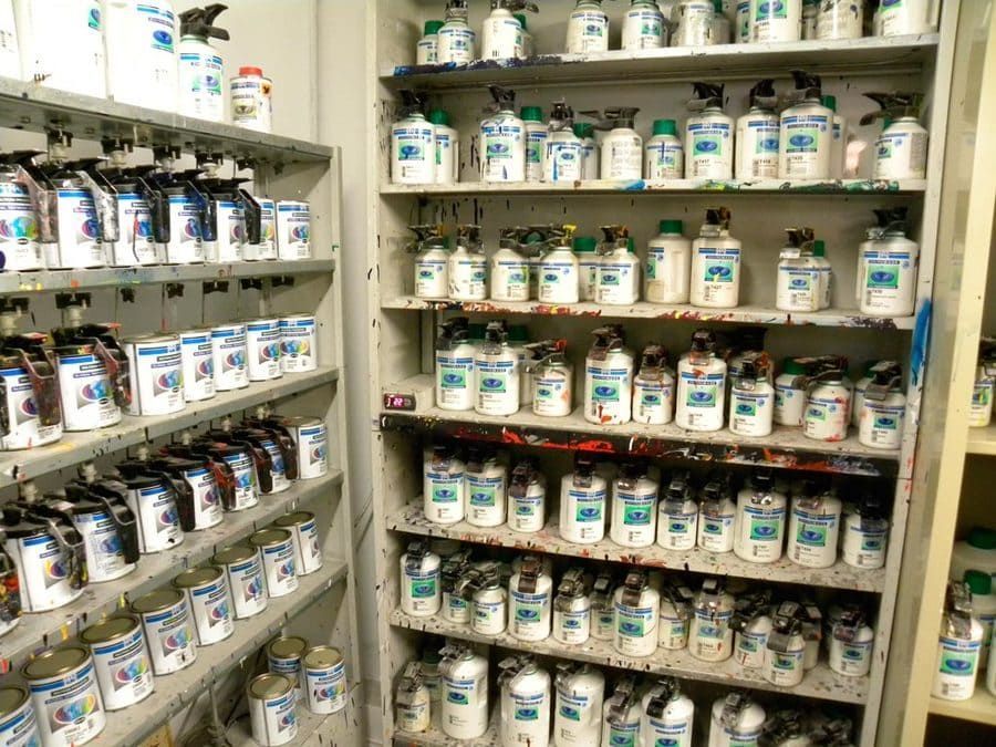 Estantes llenos de latas de pintura de distintos tamaños, en su mayoría blancas con etiquetas azules y verdes, en una tienda de suministros de pintura.
