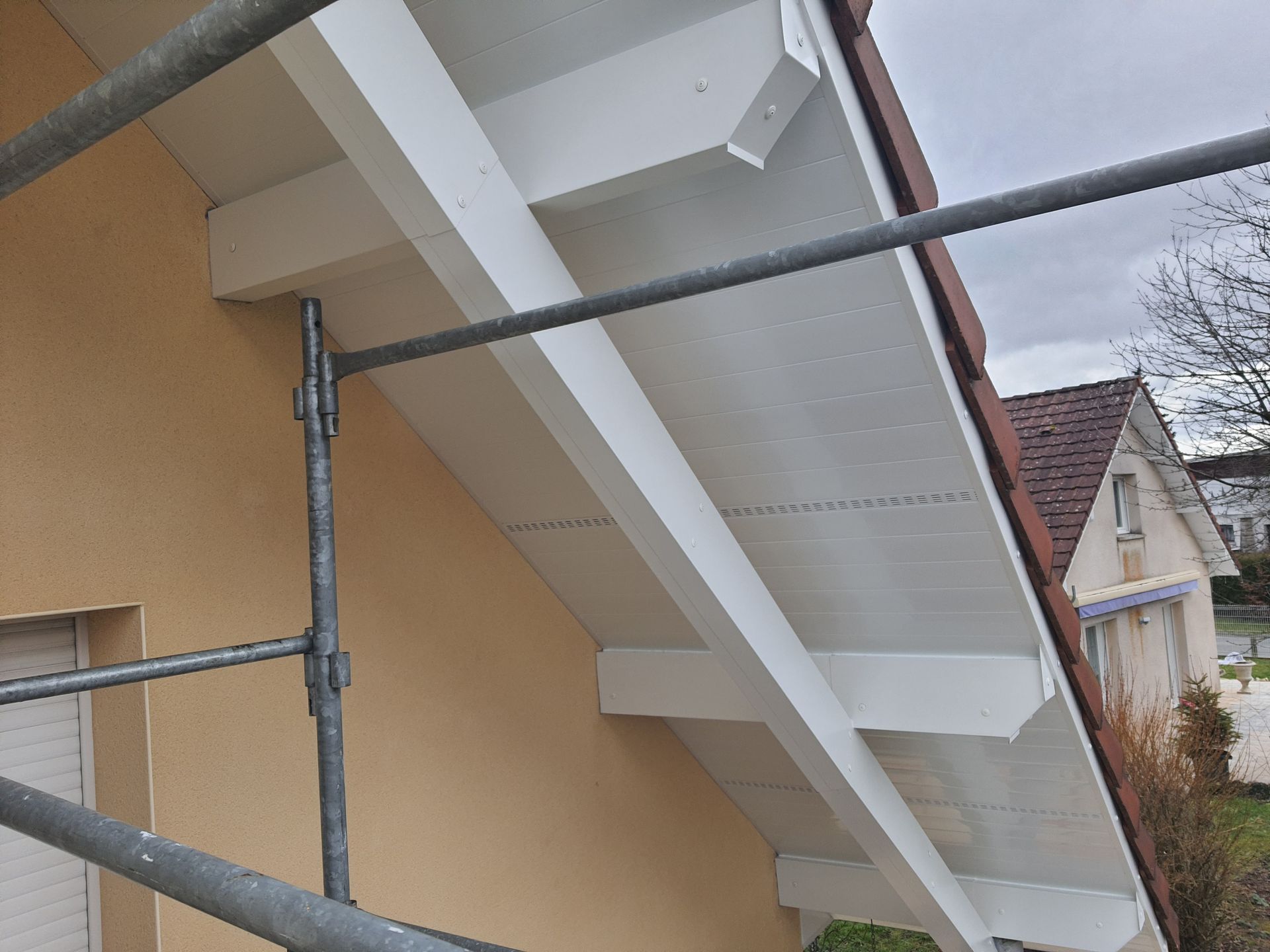 Poutres peintes en blanc sous un avant-toit, soutenues par un échafaudage sur un mur beige.