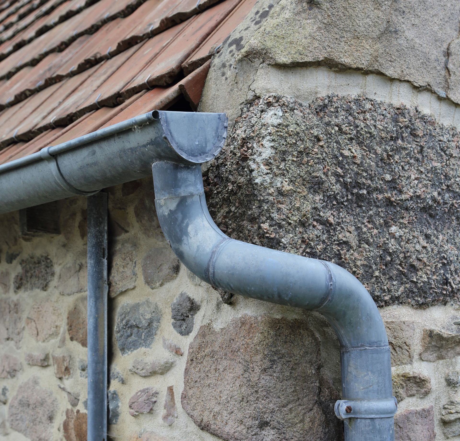 Gouttière en métal gris sur un bâtiment en pierre avec un toit de tuiles rouges.