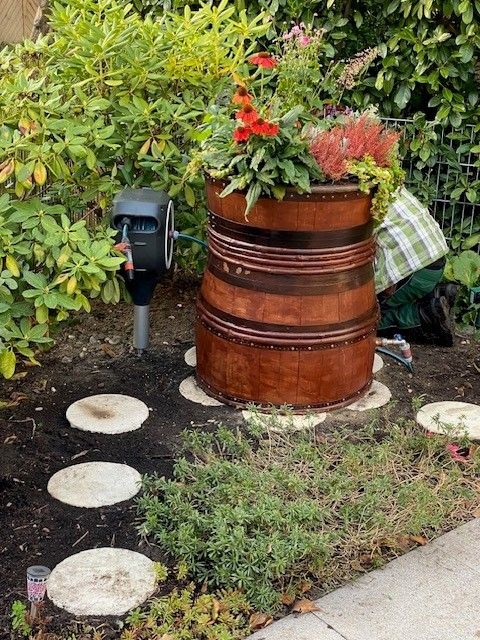 Mitten in einem Garten steht ein mit Blumen gefülltes Holzfass.
