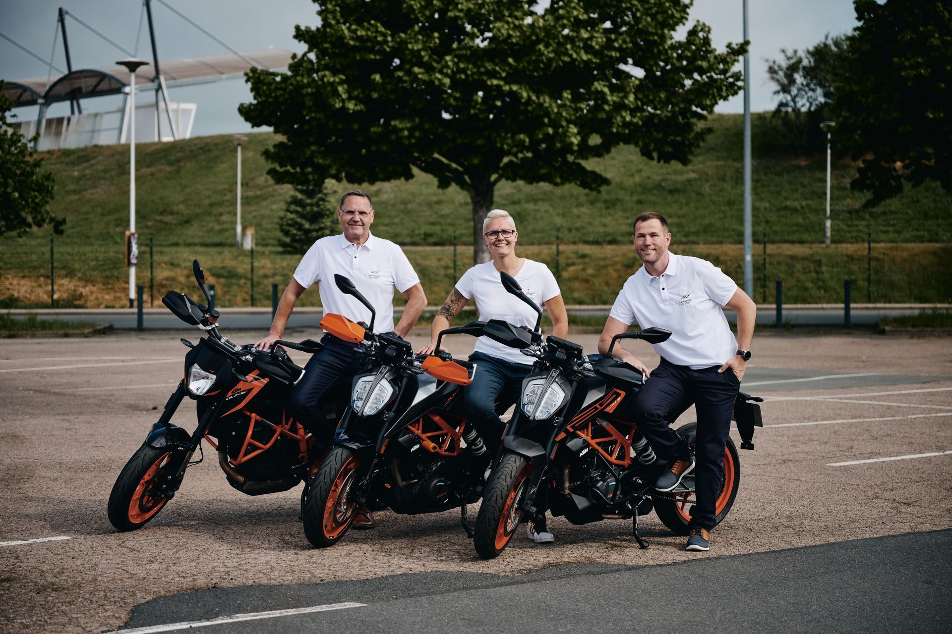 Drei lächelnde Männer neben orangefarbenen und schwarzen Motorrädern auf einem Parkplatz im Freien.