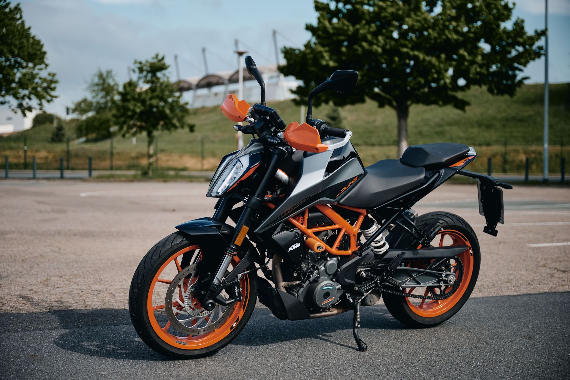 Schwarz-orangefarbenes KTM Duke-Motorrad auf Asphalt geparkt. Bäume und ein verschwommenes Gebäude im Hintergrund.