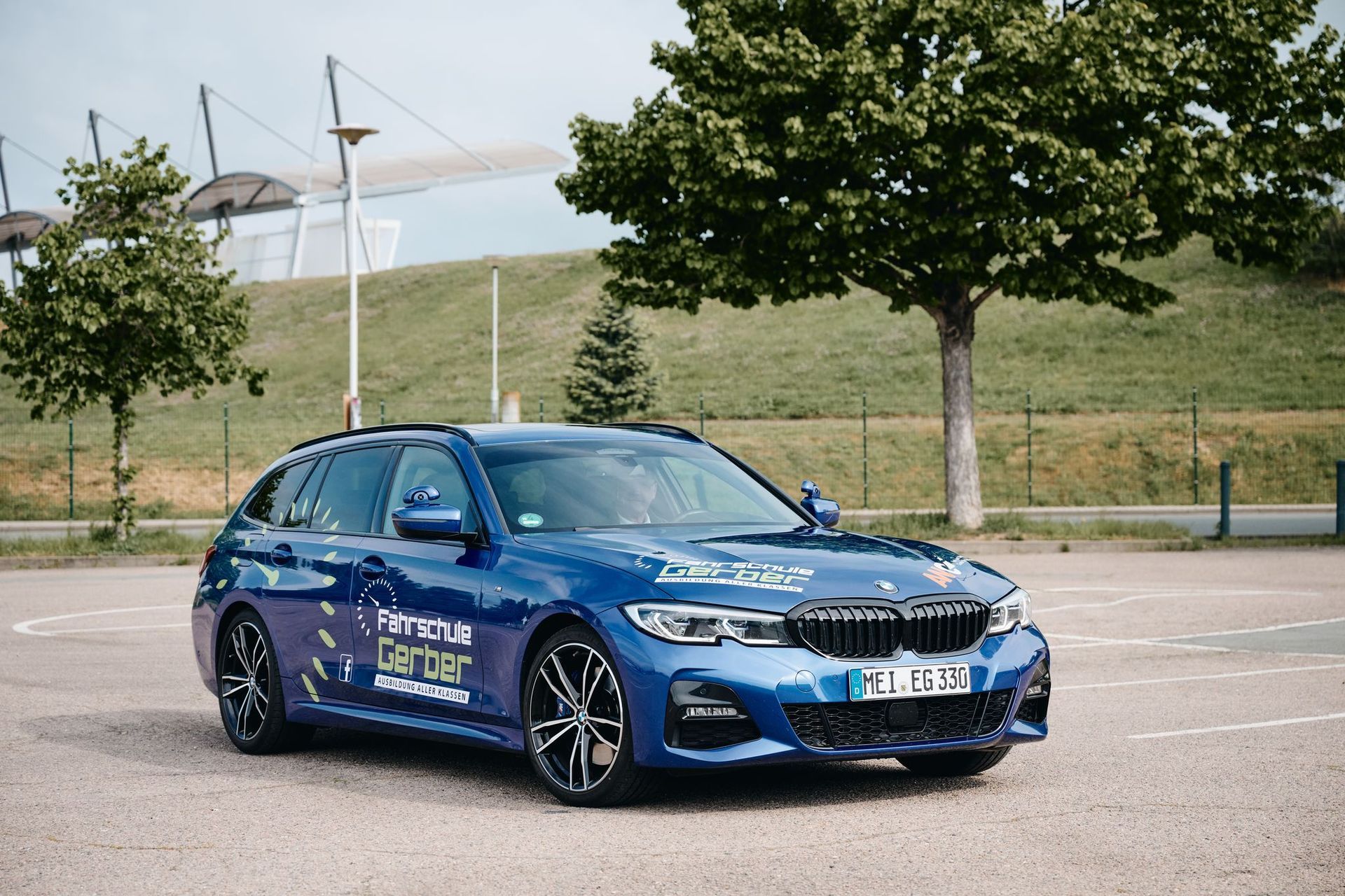Draußen parkt ein blauer BMW-Kombi; im Hintergrund sind Bäume und ein Gebäude zu sehen.