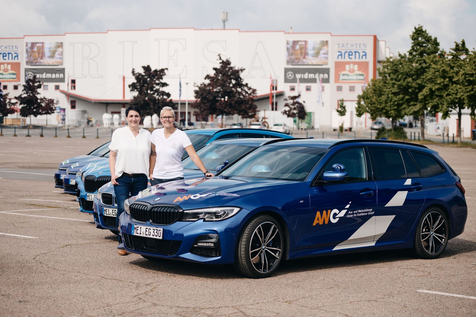 Zwei Personen stehen neben blauen Autos auf einem Parkplatz vor einer Sportarena.