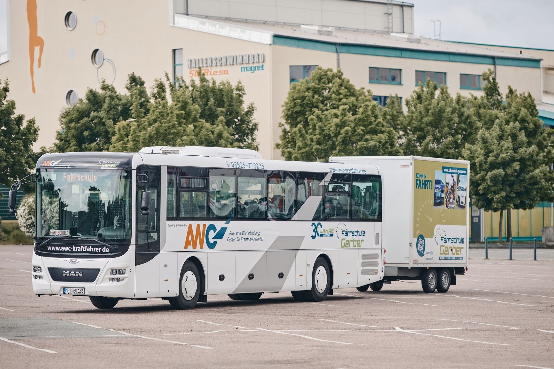 Weißer Bus mit Anhänger und „ANC“-Logo, geparkt vor einem Gebäude mit Bäumen.