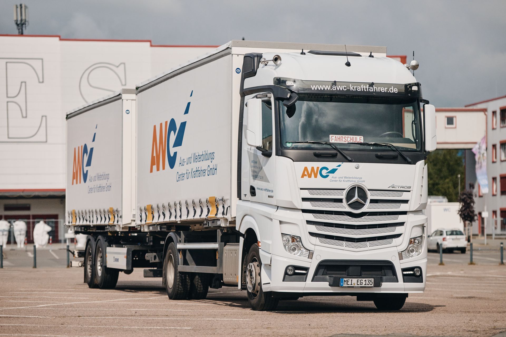 Weißer ANC-Lieferwagen auf einem Parkplatz, mit einem Mercedes-Benz-Logo auf der Vorderseite und einem großen Lagerhaus im Hintergrund.