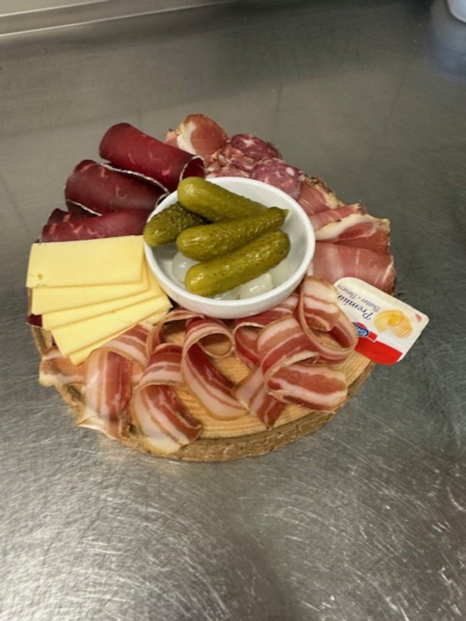 Planche de charcuterie avec cornichons, fromage, viande et beurre sur une surface métallique.
