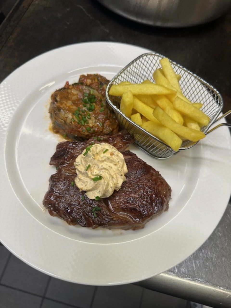 Steak garni de beurre, frites dans un panier métallique et viande braisée sur une assiette blanche.