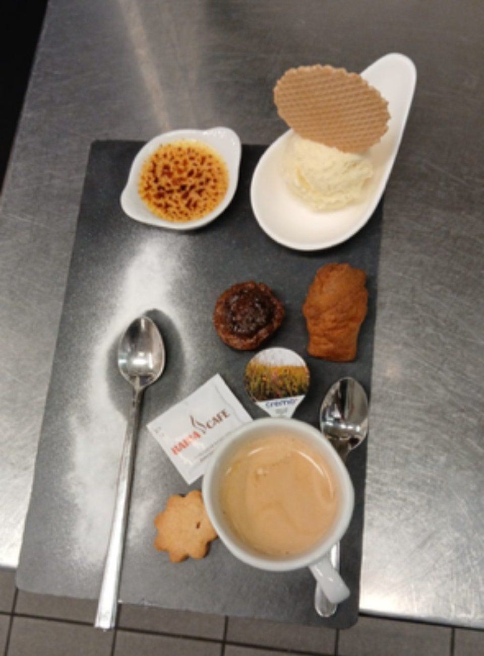 Assiette à dessert : crème brûlée, glace, viennoiseries, café, cuillère, sachet de sucre sur une surface métallique.