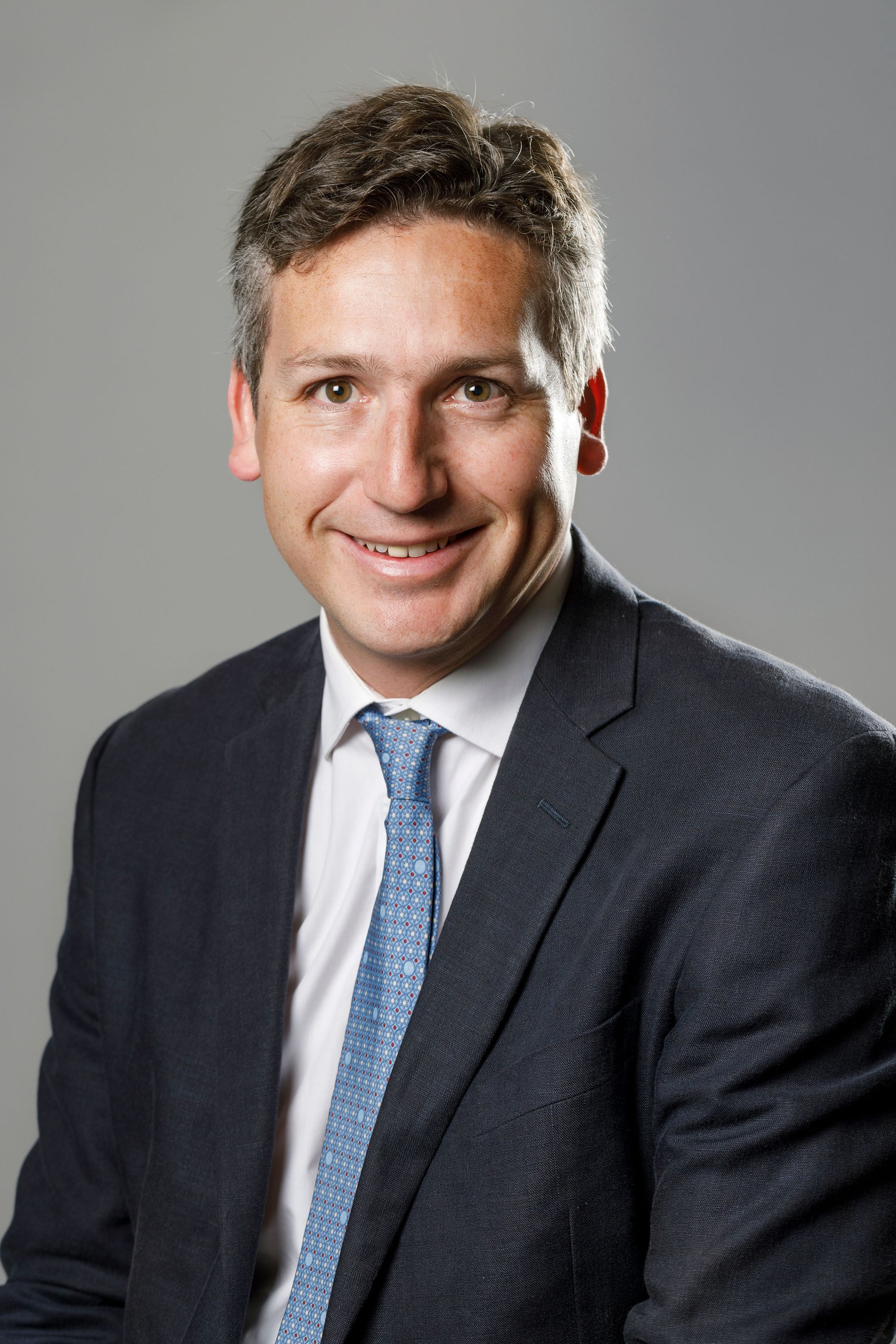 Hombre de traje sonriendo, mirando hacia adelante; piel clara, cabello oscuro, corbata azul.