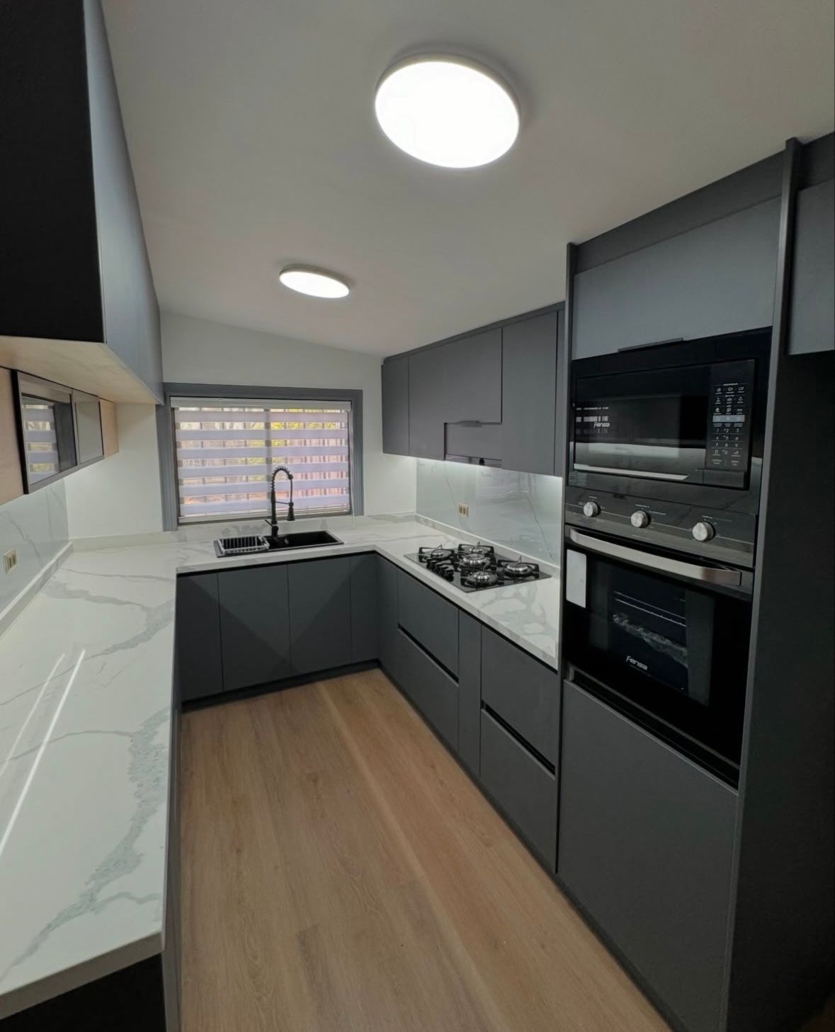 Cocina moderna de color gris con encimeras blancas y suelos de madera, equipada con horno y microondas empotrados.