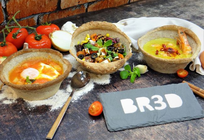 Três tigelas de comida repletas de pratos diferentes, sobre uma mesa rústica com tomates e uma placa com a inscrição “BR3D”.