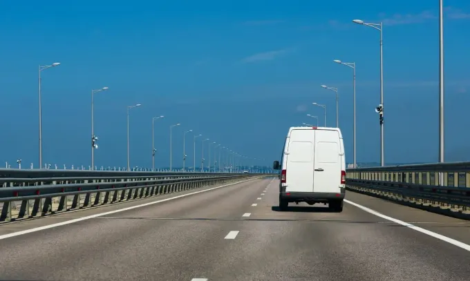 Una furgoneta blanca se aleja por una autopista abierta de varios carriles bajo un cielo azul despejado.