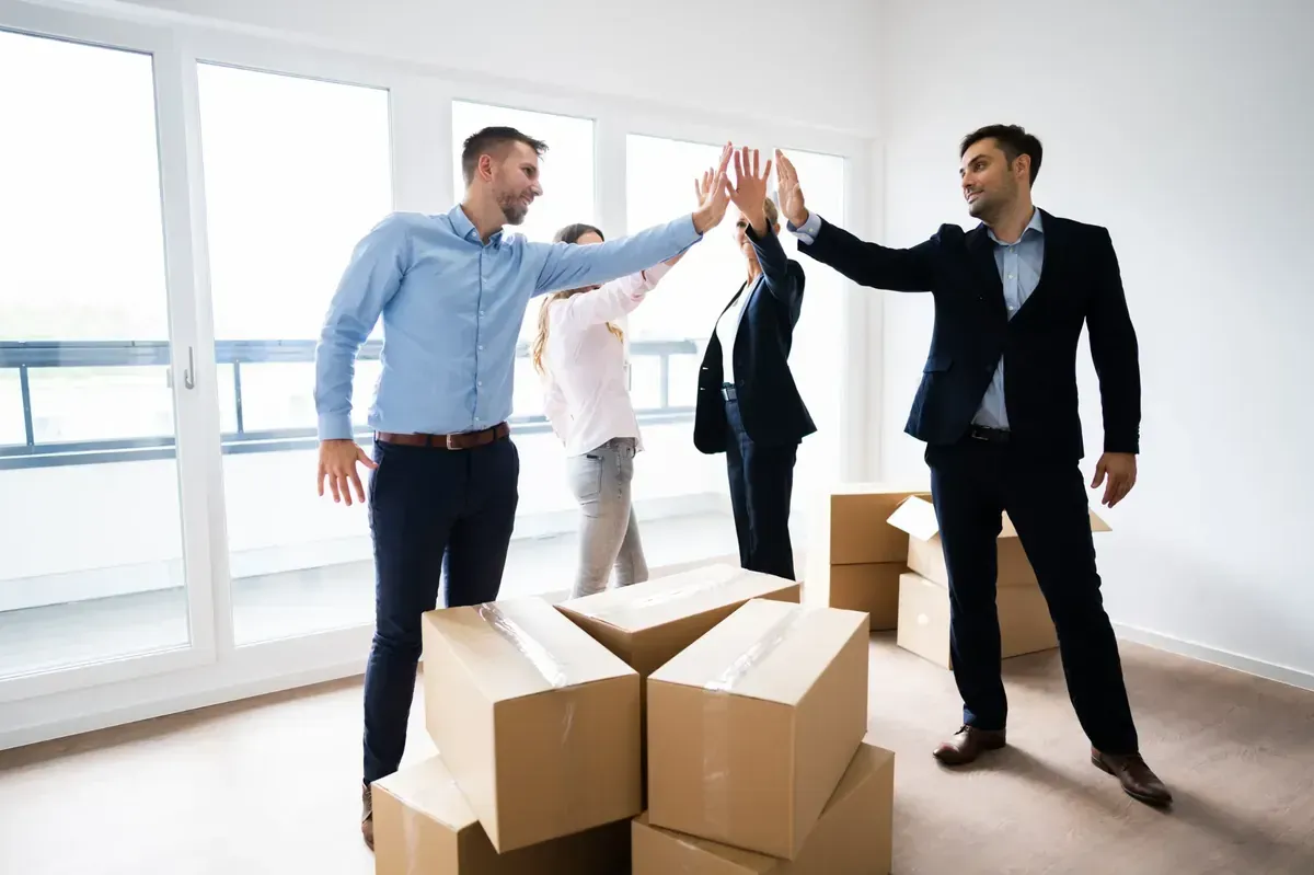 Cuatro personas en una habitación luminosa y vacía, con cajas de cartón de mudanza, chocan las manos para celebrar.