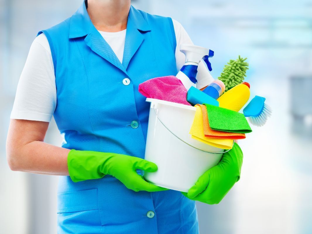 Una mujer que lleva guantes verdes sostiene un balde con productos de limpieza.
