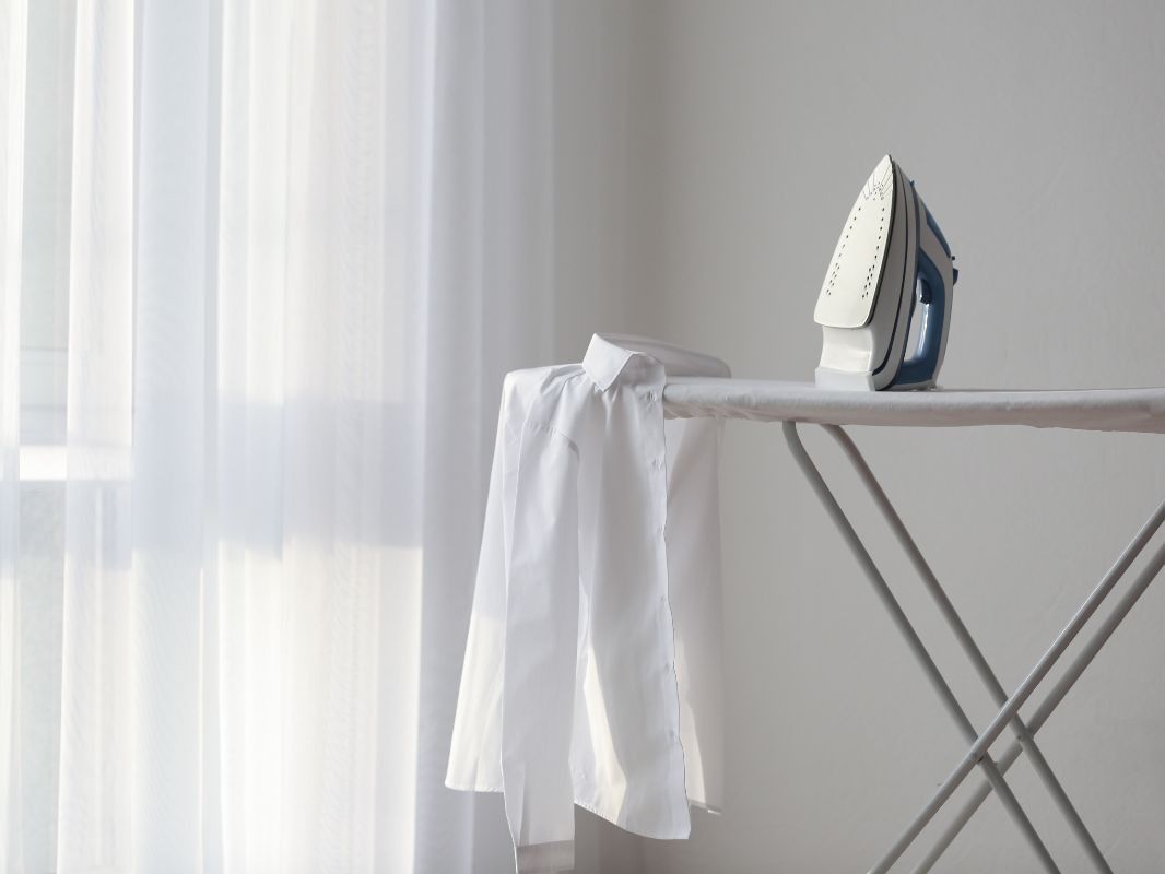 Una camisa blanca está colgada en una tabla de planchar al lado de una ventana.