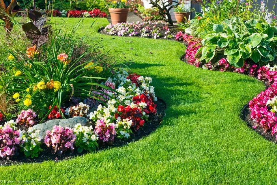 Bunte Blumenbeete säumen einen üppig grünen Gartenweg mit Topfpflanzen und Sträuchern.
